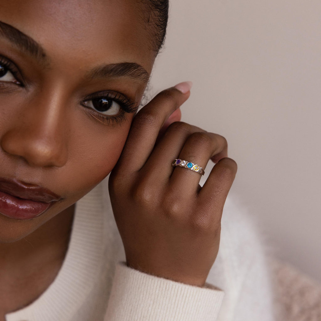 A woman with natural makeup displays the Vintage Multiple Birthstone Ring, featuring a gold band set with colorful gemstones on her finger.