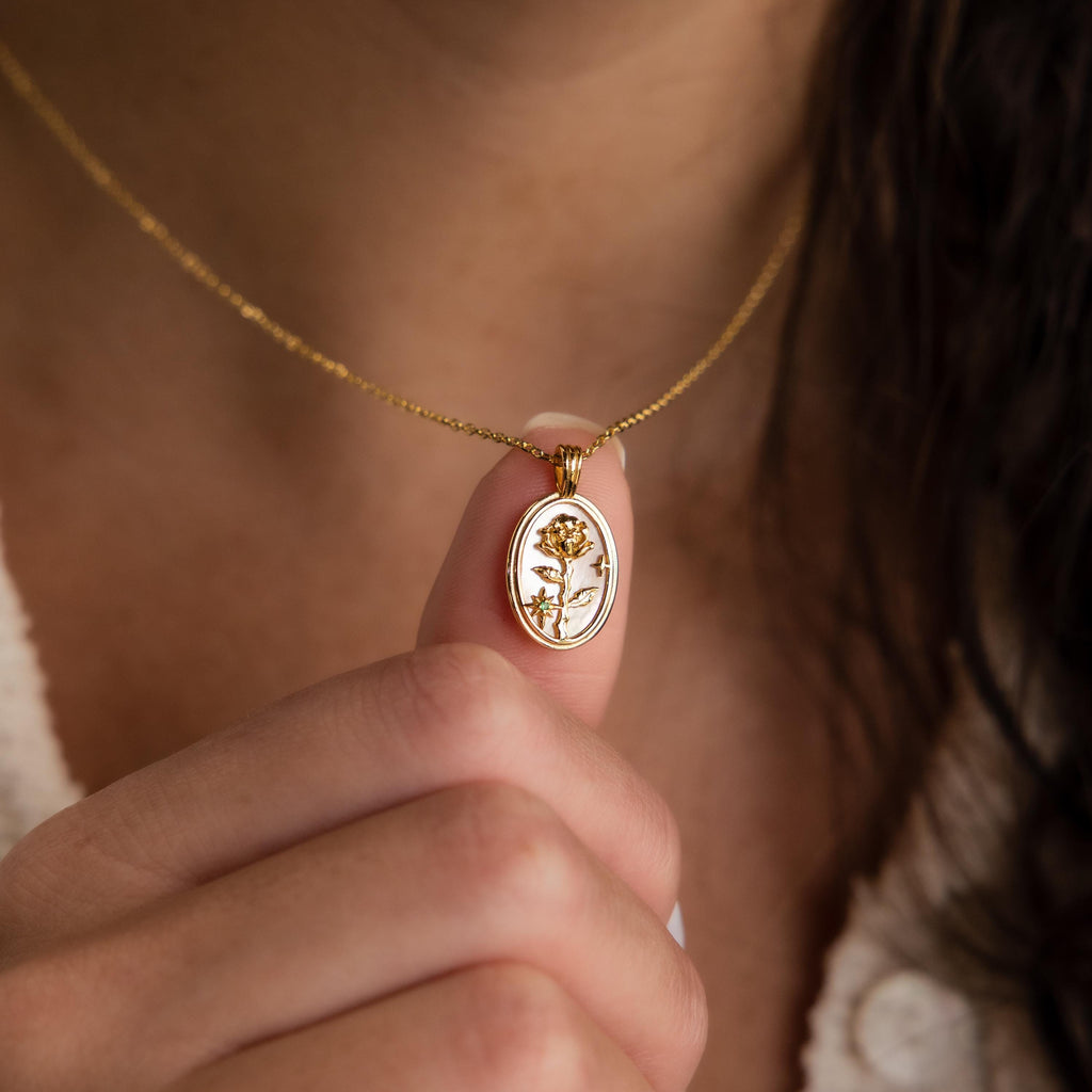 A person holds the Pearl Flower Birthstone Necklace—a gold oval mother of pearl pendant with an embossed flower—close to their chest. The pendant is suspended from a delicate 925 sterling silver chain.