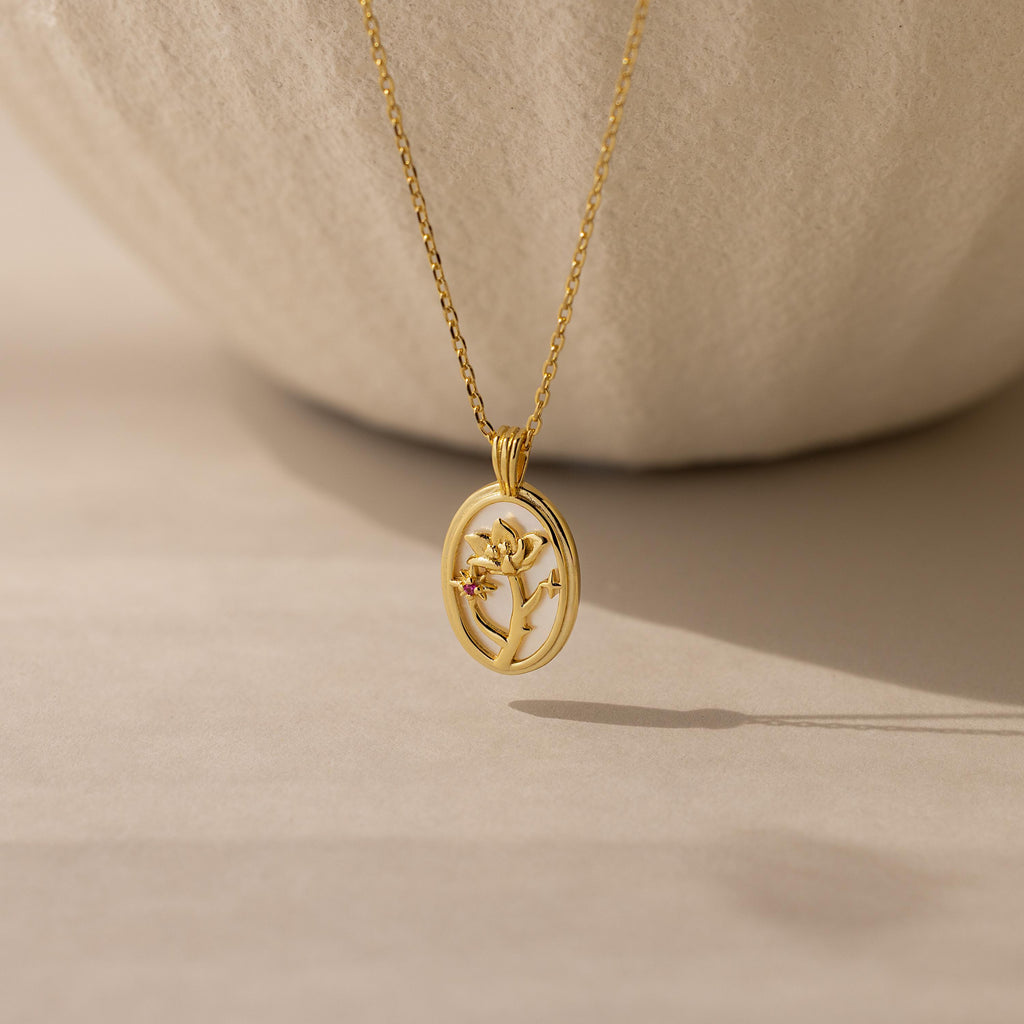 The Pearl Flower Birthstone Necklace features a gold oval mother of pearl pendant with a delicate floral design, photographed on a softly-lit neutral background with a gentle shadow beneath it.