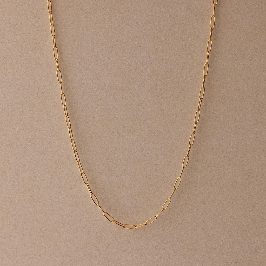 The Paperclip Chain Necklace is a delicate gold chain, ideal for layering and perfect for any style, shown on a beige background.