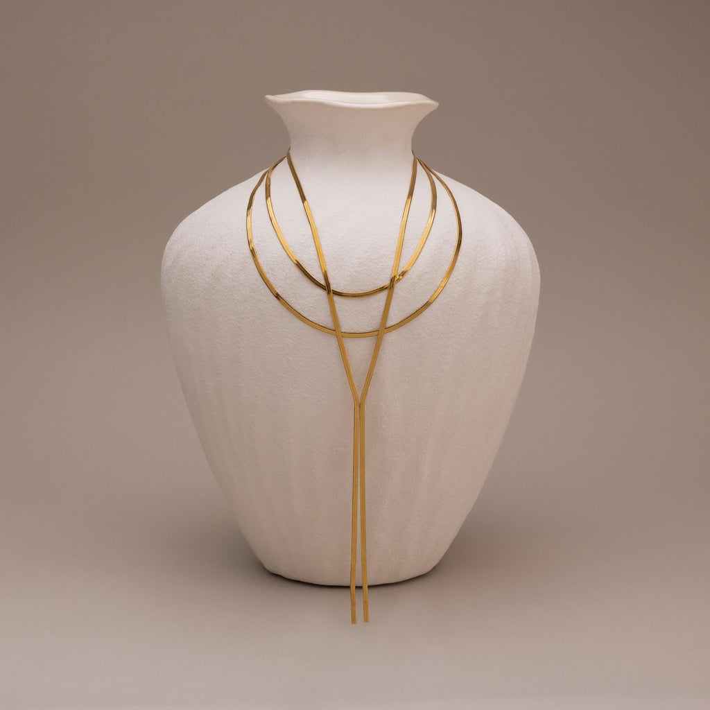 The Long Y Herringbone Chain Necklace is displayed draped over a white textured vase, set against a neutral background.