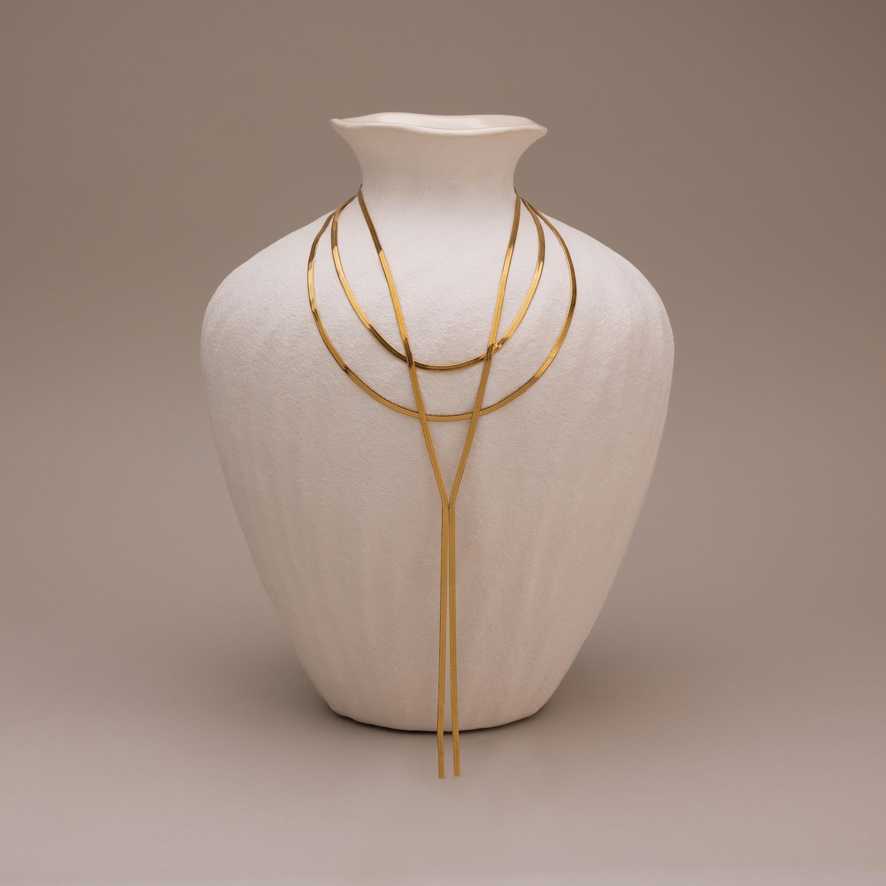 The Long Y Herringbone Chain Necklace is displayed draped over a white textured vase, set against a neutral background.