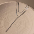 A Long Y Herringbone Chain Necklace in thin silver is displayed in a branching pattern on a round, textured beige ceramic dish.