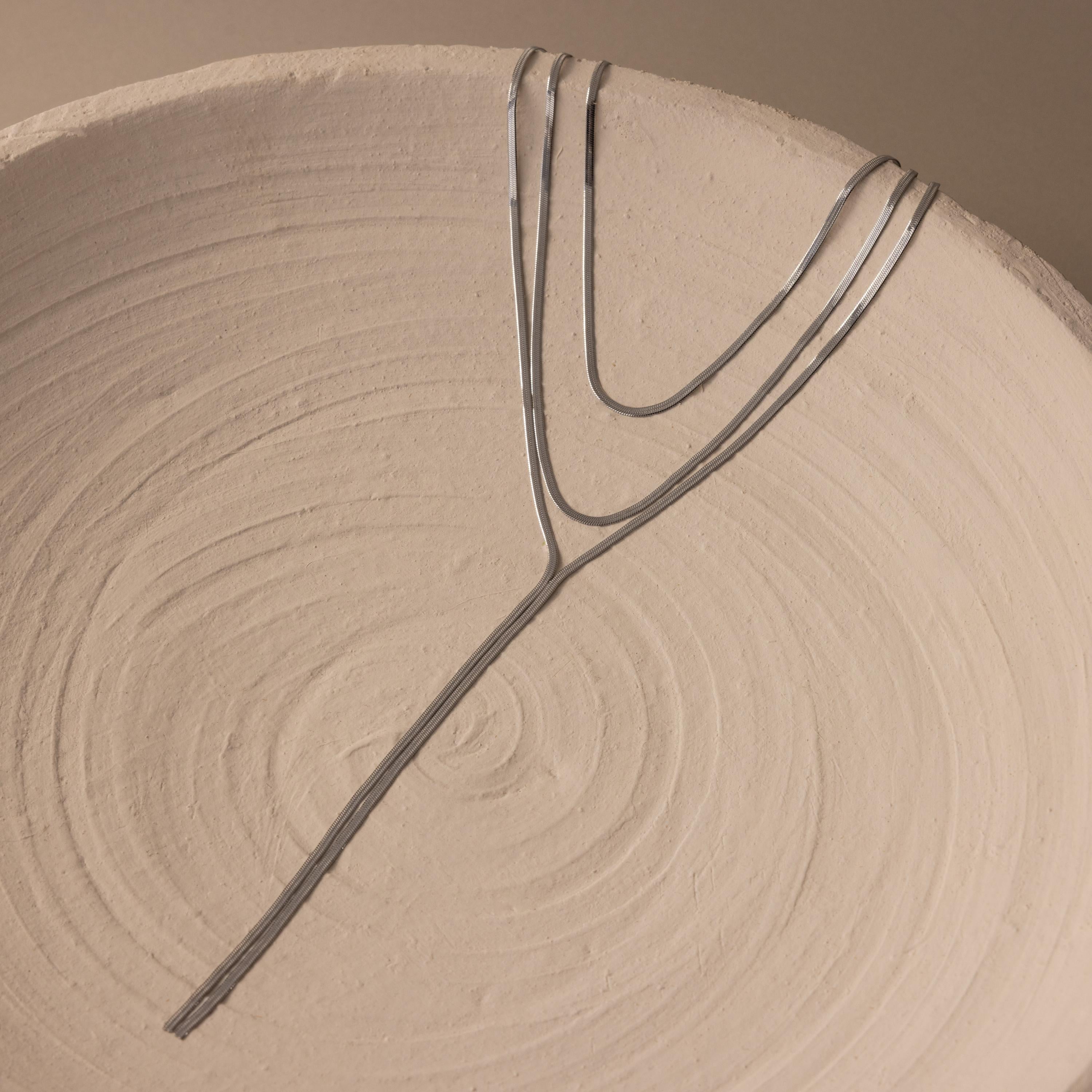 A Long Y Herringbone Chain Necklace in thin silver is displayed in a branching pattern on a round, textured beige ceramic dish.
