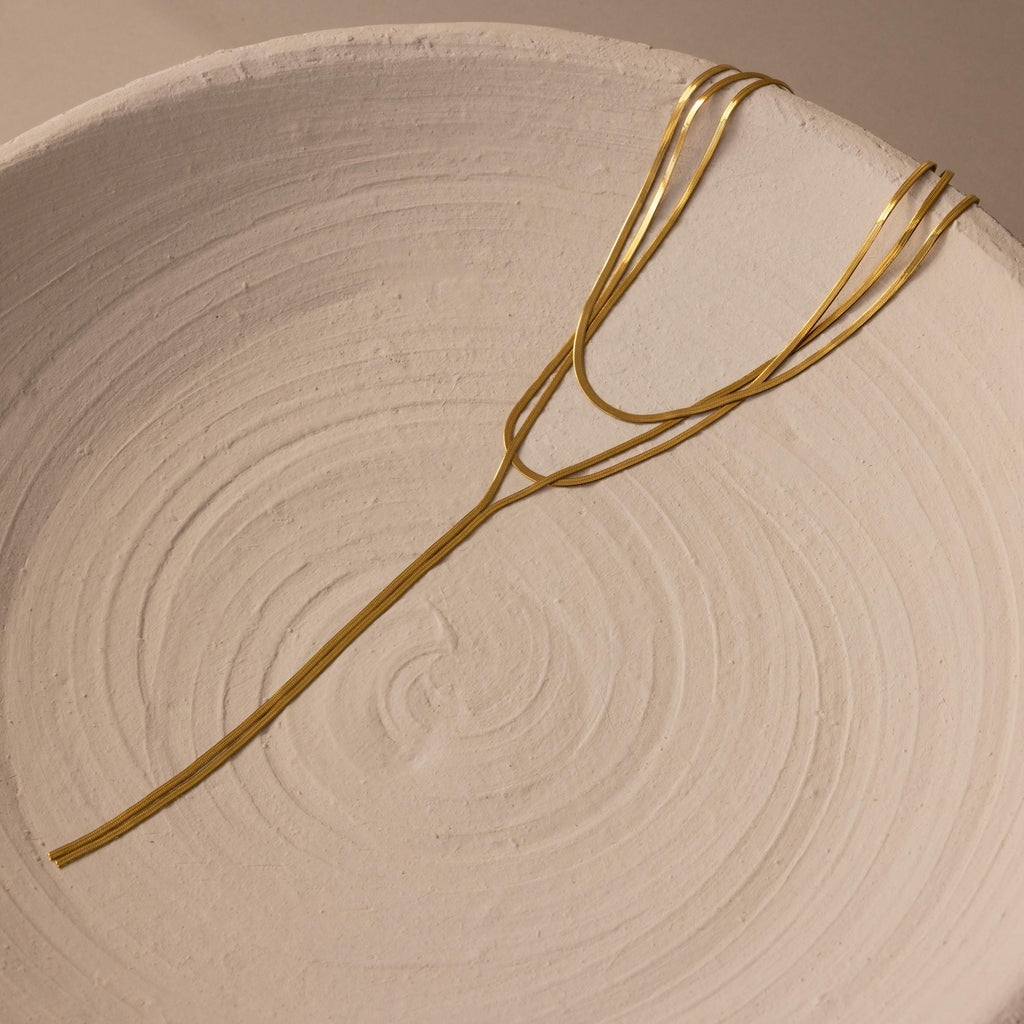 The Long Y Herringbone Chain Necklace is displayed on the edge of a textured, off-white ceramic bowl.