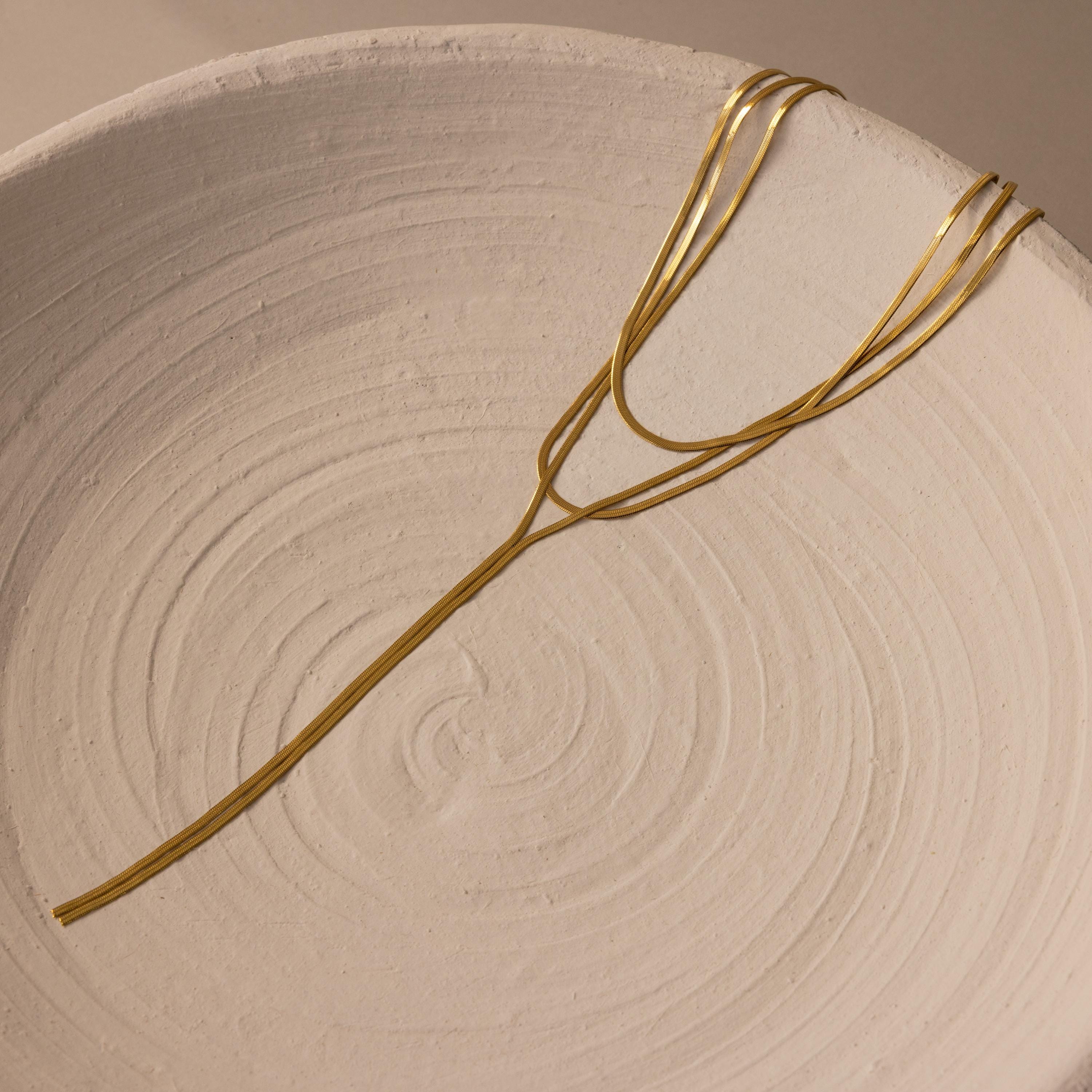 The Long Y Herringbone Chain Necklace is displayed on the edge of a textured, off-white ceramic bowl.