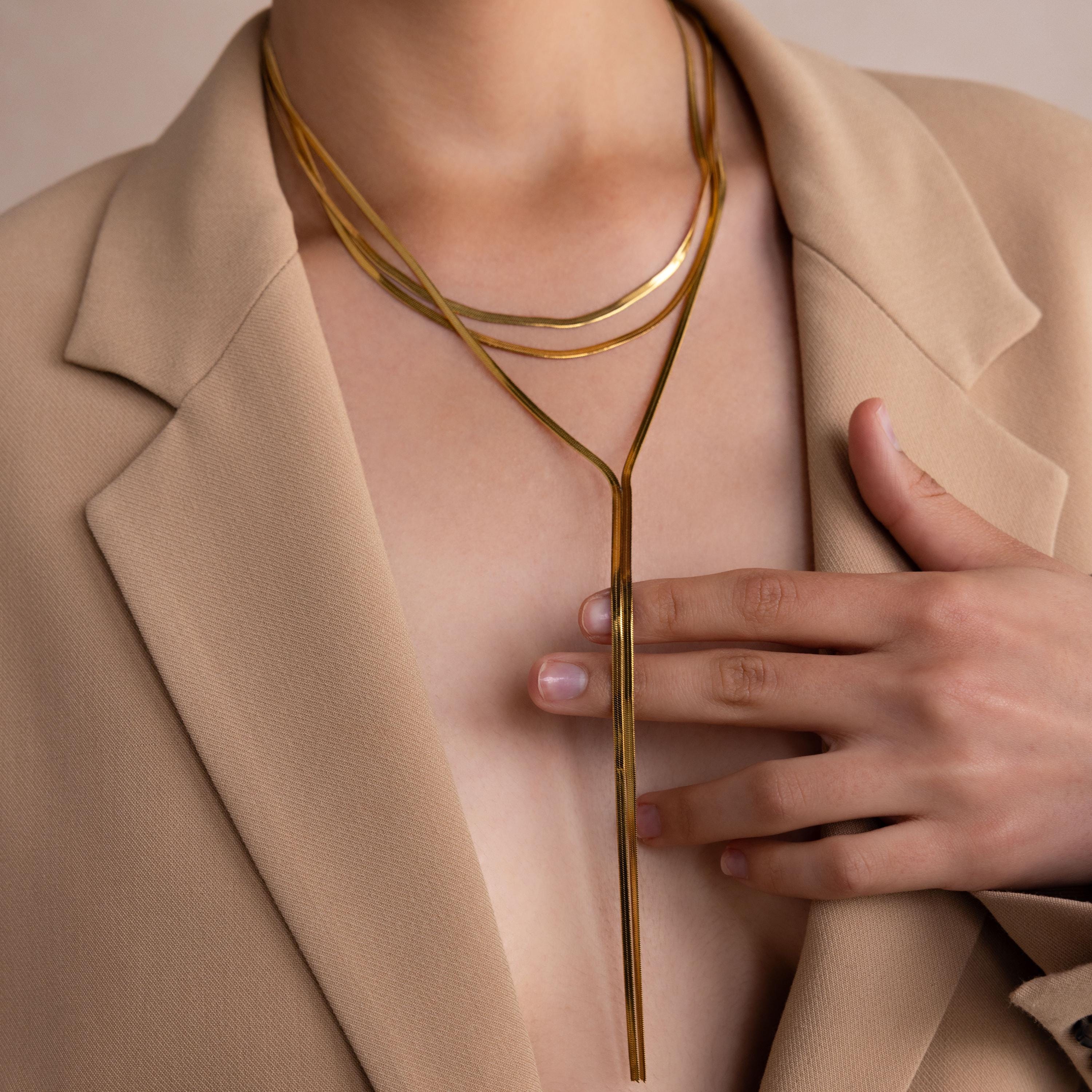 A person in a beige blazer wears layered gold necklaces, featuring the Long Y Herringbone Chain Necklace, with their hand resting on their chest.