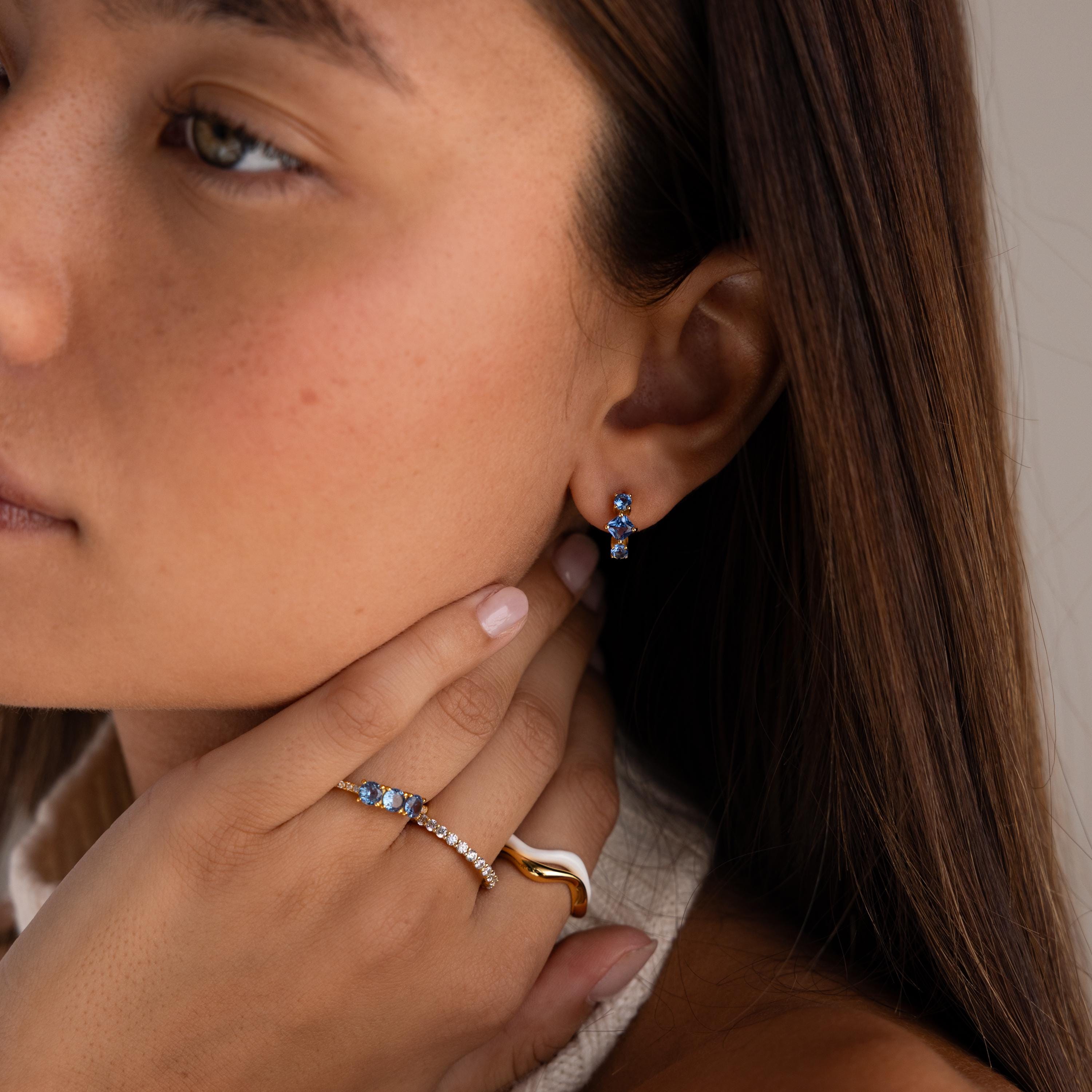 Woman wearing Princess Cut Birthstone Huggies earrings and rings, touching her neck with one hand, close-up side view.