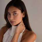 A woman wearing a sleeveless knit top and gold Princess Cut Birthstone Huggies looks at the camera, her hand near her neck.