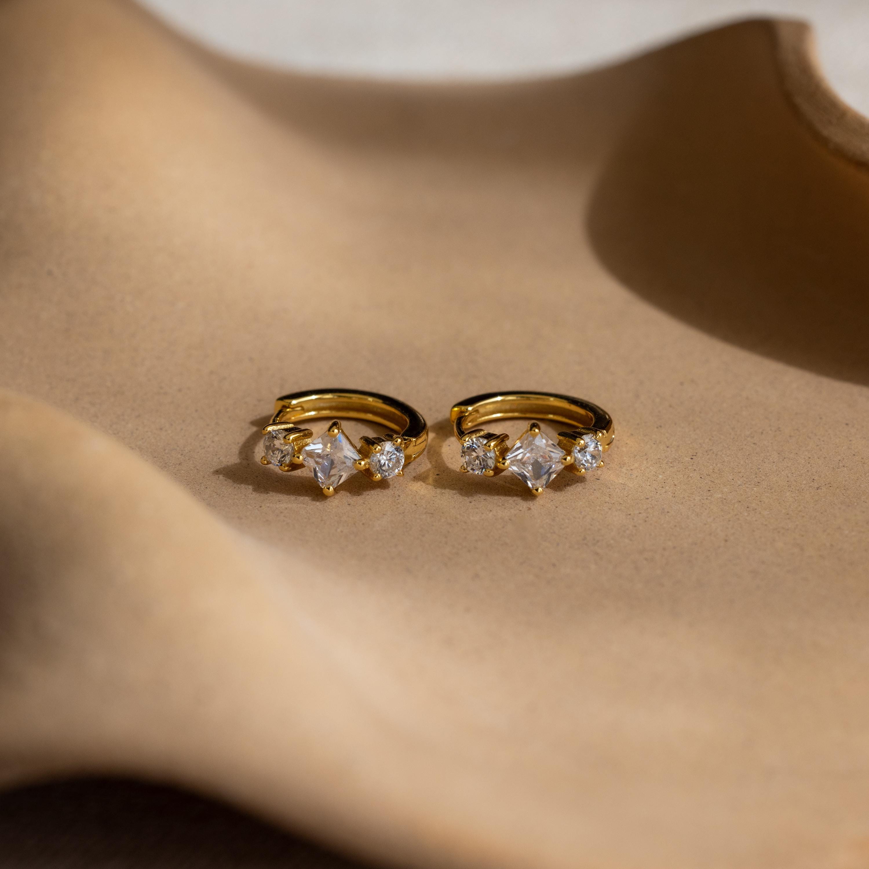 Princess Cut Birthstone Huggies are shown on a beige, wavy surface under soft lighting.