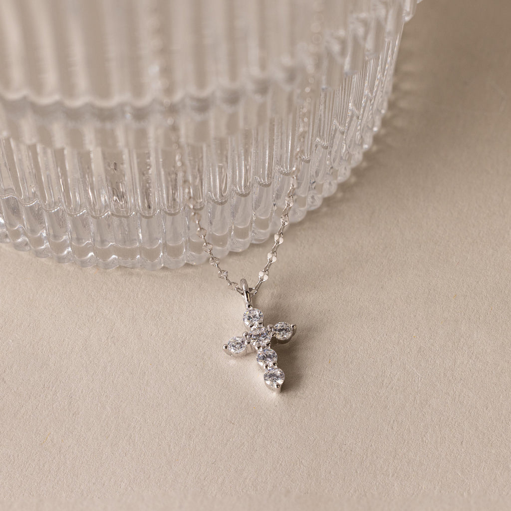 The Diamond Cross Necklace, featuring a silver chain and a small diamond cross pendant accented with pave CZ stones, is displayed beside a ribbed glass container on a beige surface.