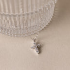 The Diamond Cross Necklace, featuring a silver chain and a small diamond cross pendant accented with pave CZ stones, is displayed beside a ribbed glass container on a beige surface.