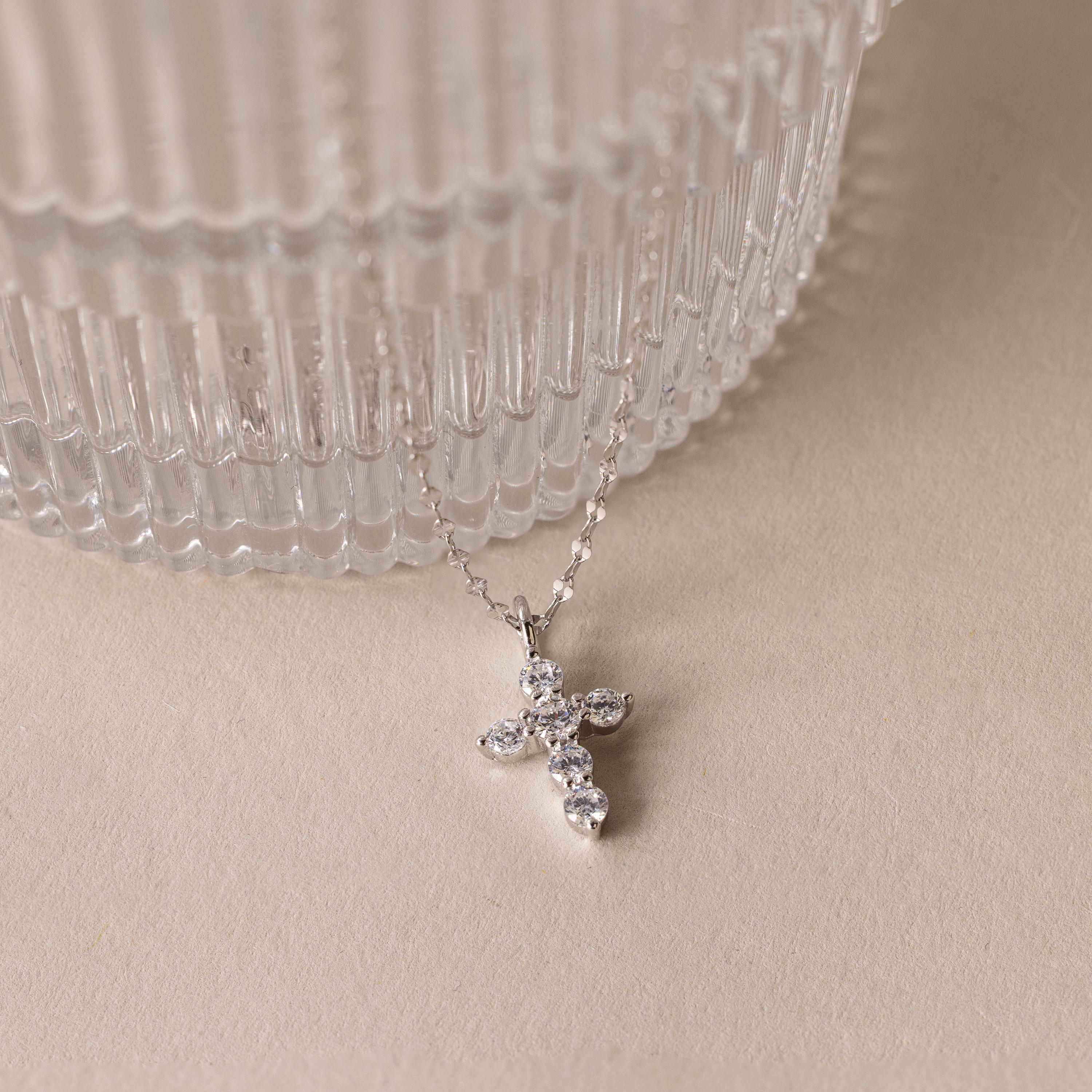 The Diamond Cross Necklace, featuring a silver chain and a small diamond cross pendant accented with pave CZ stones, is displayed beside a ribbed glass container on a beige surface.
