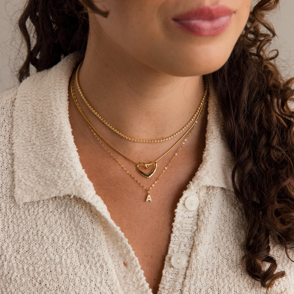 Woman wearing a cream textured shirt and layered gold necklaces, including the Dainty Pave Letter Necklace in 18K Gold with CZ diamonds and a delicate heart pendant.