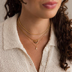 Woman wearing a cream textured shirt and layered gold necklaces, including the Dainty Pave Letter Necklace in 18K Gold with CZ diamonds and a delicate heart pendant.
