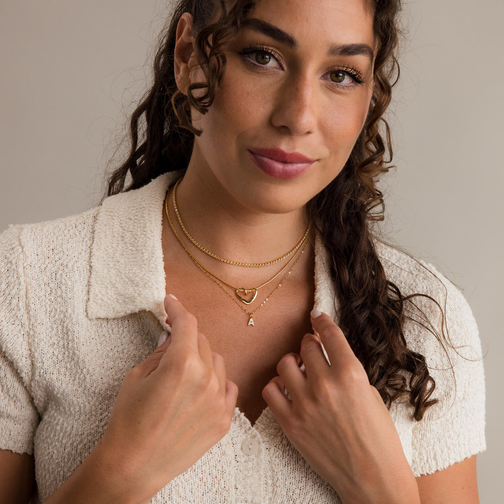 A woman with curly hair wears a cream textured collared shirt and the Dainty Pave Letter Necklace in 18K Gold, layered with other gold necklaces, as she looks at the camera.
