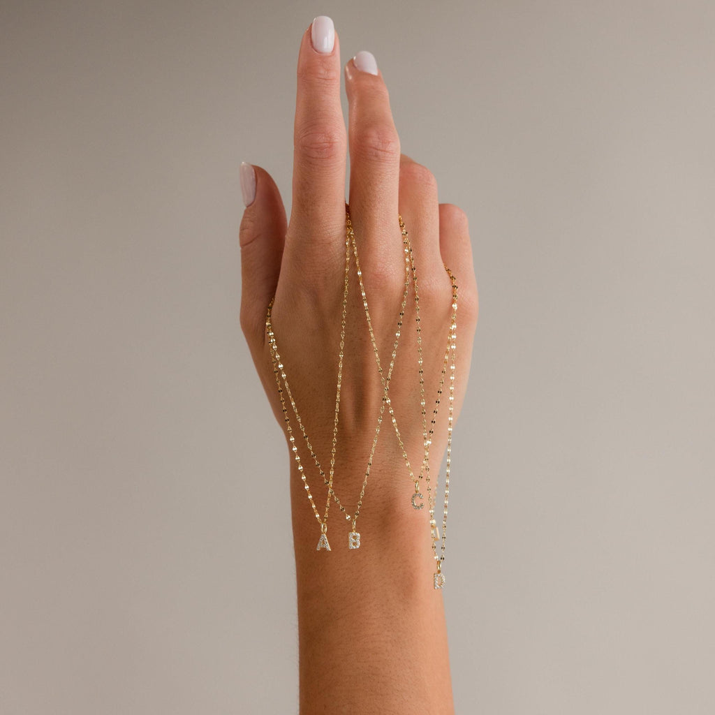 A hand displays four Dainty Pave Letter Necklaces in 18K Gold, each featuring a sparkling CZ diamond letter charm—A, B, C, and R—on delicate mirror chains.