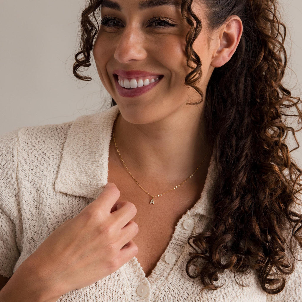 Smiling woman with curly hair wears a cream knit top and the Dainty Pave Letter Necklace, featuring a gold A pendant.