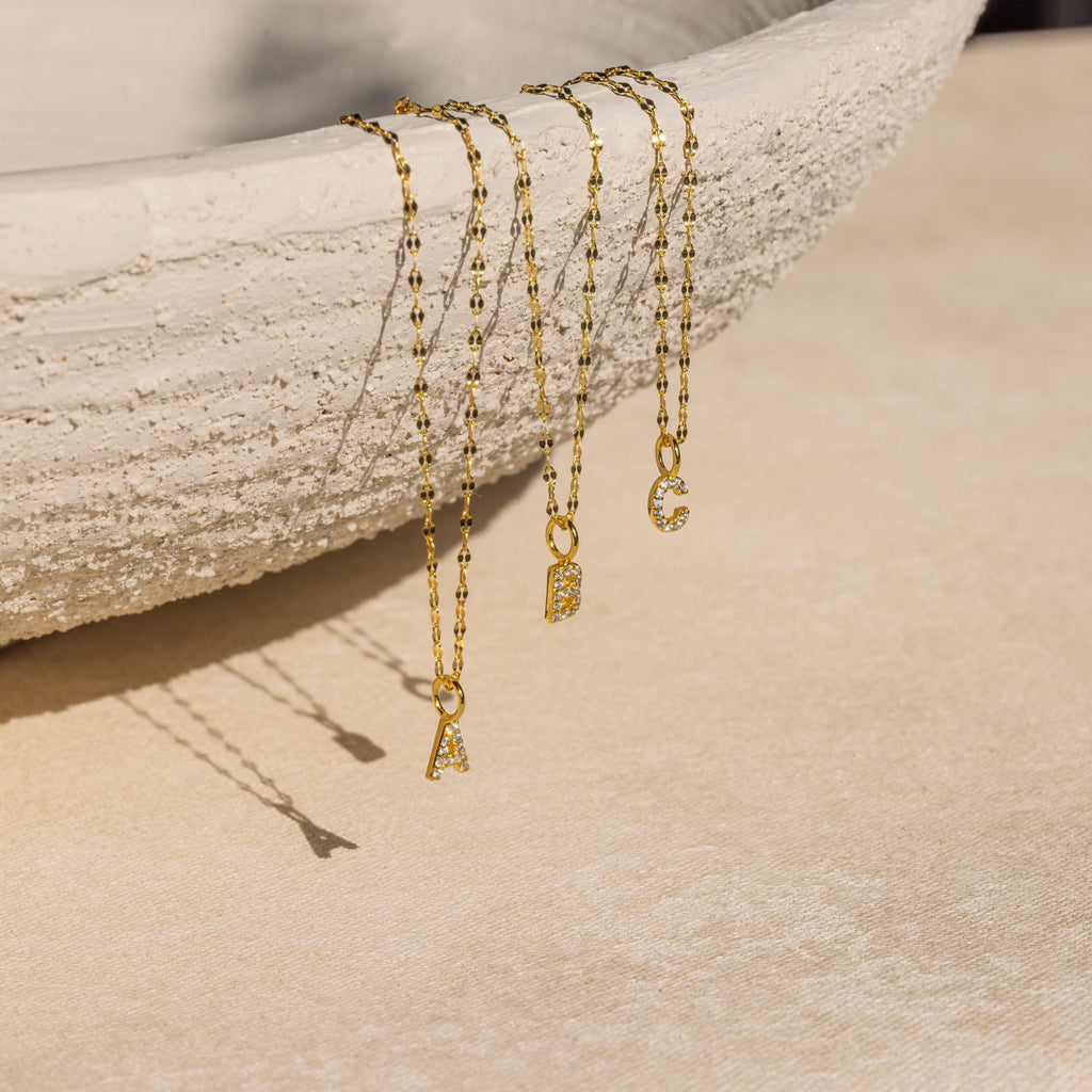 Three Dainty Pave Letter Necklaces in 18K Gold with sparkling CZ diamonds, featuring A, B, and C pendants, drape over the edge of a textured beige bowl.