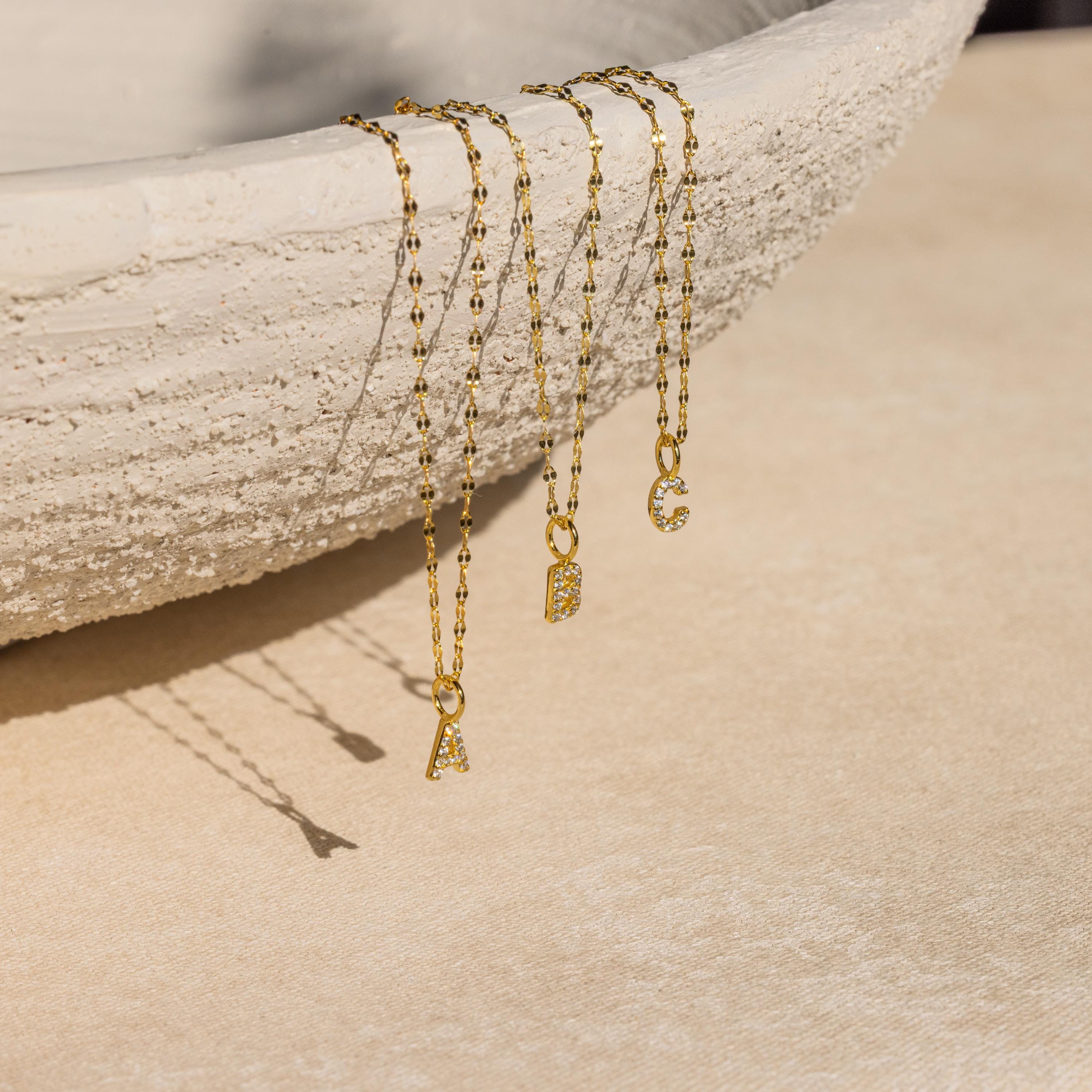 Three Dainty Pave Letter Necklaces in 18K Gold with sparkling CZ diamonds, featuring A, B, and C pendants, drape over the edge of a textured beige bowl.