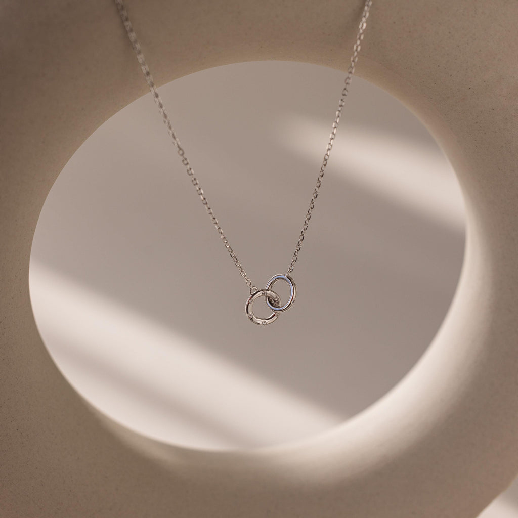 Pave Interlocking Circle Necklace in silver, featuring two intertwined rings symbolizing love, shown on a soft beige circular background with gentle shadows.
