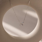 Pave Interlocking Circle Necklace in silver, featuring two intertwined rings symbolizing love, shown on a soft beige circular background with gentle shadows.