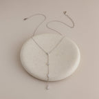 The Mixed Diamond Lariat Necklace is an elegant, delicate silver Y-shaped piece adorned with diamond gemstones, showcased on a round, light stone.