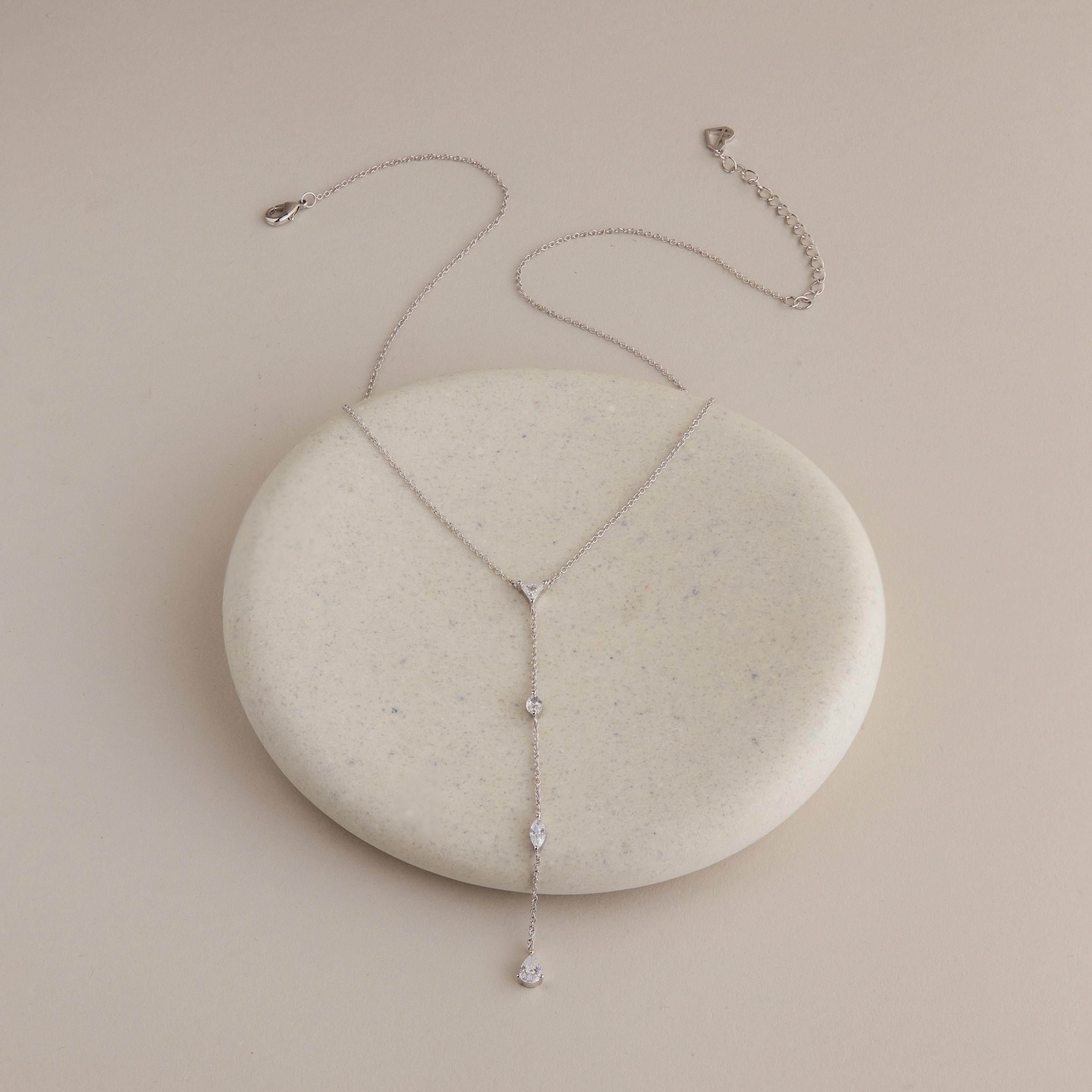 The Mixed Diamond Lariat Necklace is an elegant, delicate silver Y-shaped piece adorned with diamond gemstones, showcased on a round, light stone.