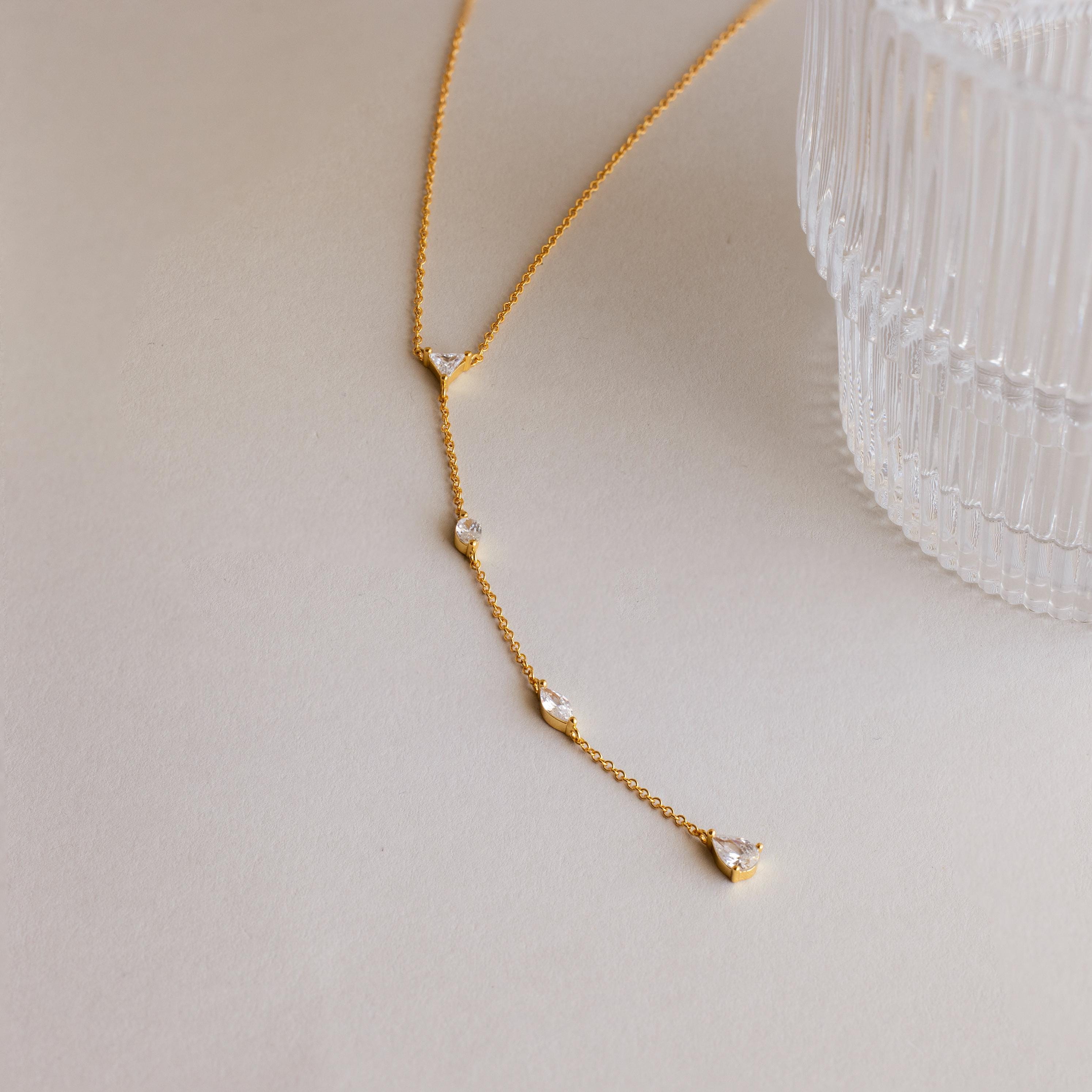 The Mixed Diamond Lariat Necklace, crafted in delicate gold and adorned with diamond gemstones, is displayed beside a clear textured glass on a light surface, highlighting the necklace's refined elegance.