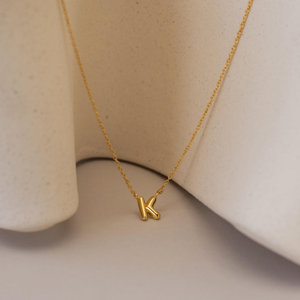 The Dainty Bubble Letter Necklace showcases a gold K bubble letter pendant, elegantly displayed on a smooth beige fabric background.