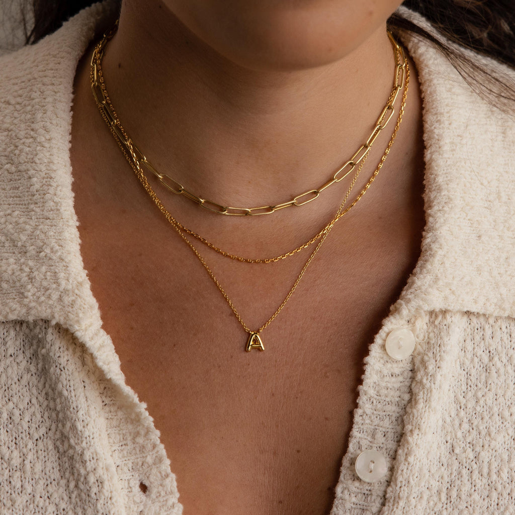 Close-up of a person layering three gold necklaces, featuring the Dainty Bubble Letter Necklace in 18K Gold with an A pendant, styled over a cream textured shirt.