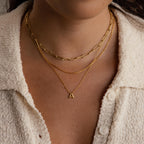 Close-up of a person layering three gold necklaces, featuring the Dainty Bubble Letter Necklace in 18K Gold with an A pendant, styled over a cream textured shirt.