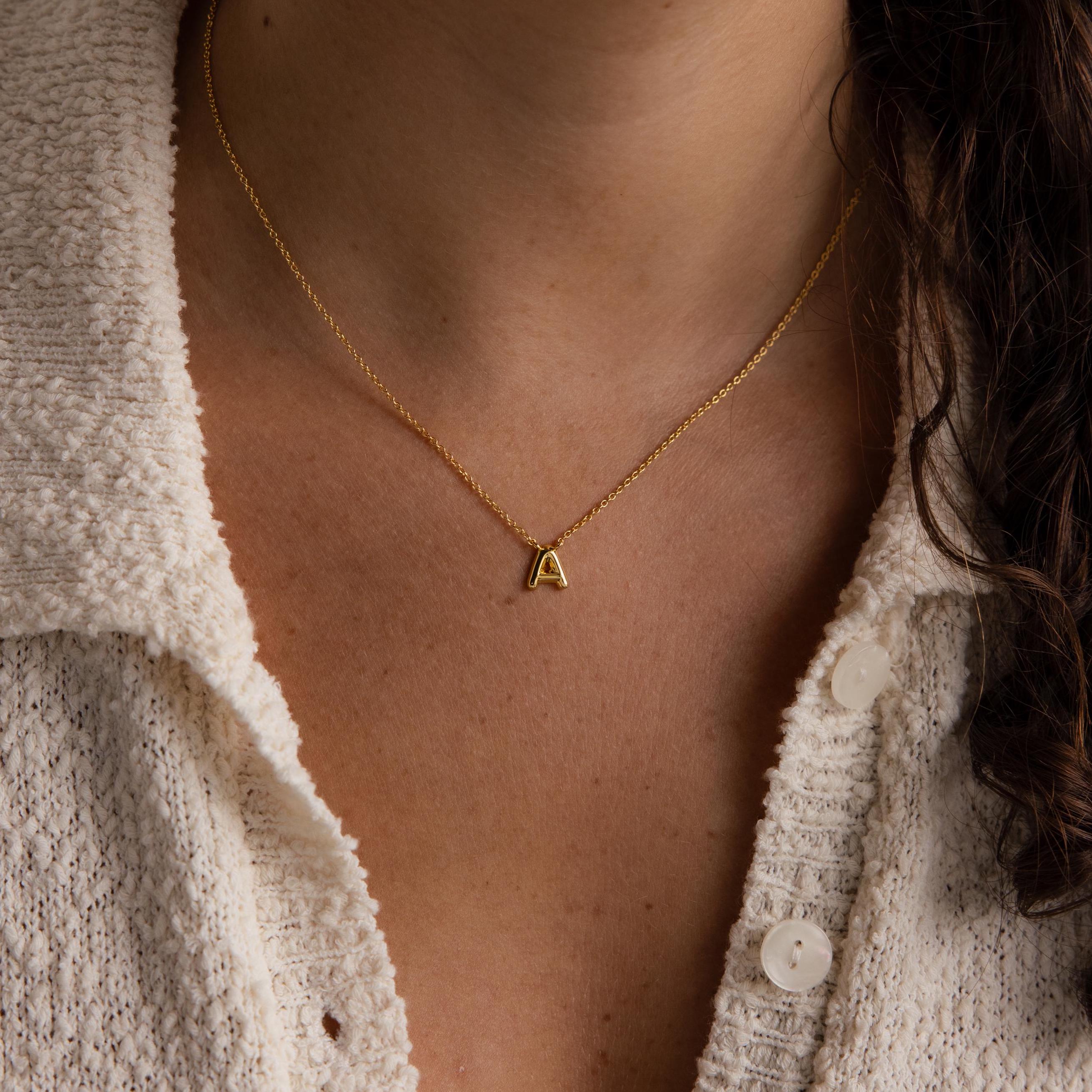 A person in a white textured shirt wears the Dainty Bubble Letter Necklace, featuring a minimalist bubble letter A pendant.