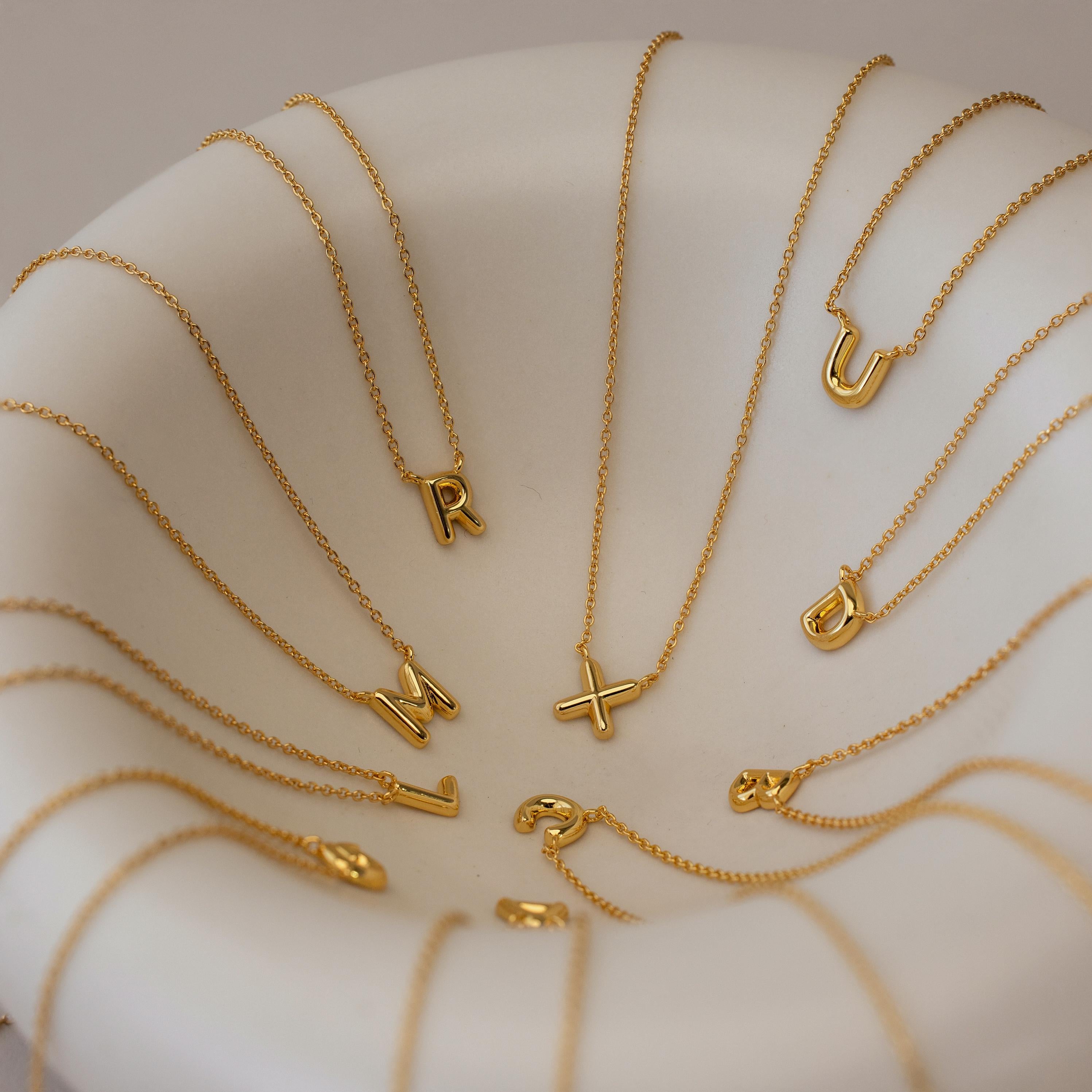 Dainty Bubble Letter Necklaces with gold letter pendants are arranged in a white bowl, displaying a variety of alphabet letters for a chic personalized necklace showcase.