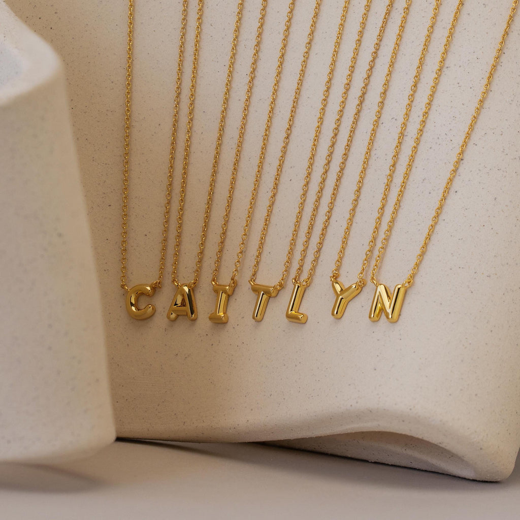 The Dainty Bubble Letter Necklace, featuring gold individual letter pendants that spell CAITLYN, is displayed on a neutral textured background for a chic and personalized accessory touch.