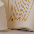 The Dainty Bubble Letter Necklace, featuring gold individual letter pendants that spell CAITLYN, is displayed on a neutral textured background for a chic and personalized accessory touch.
