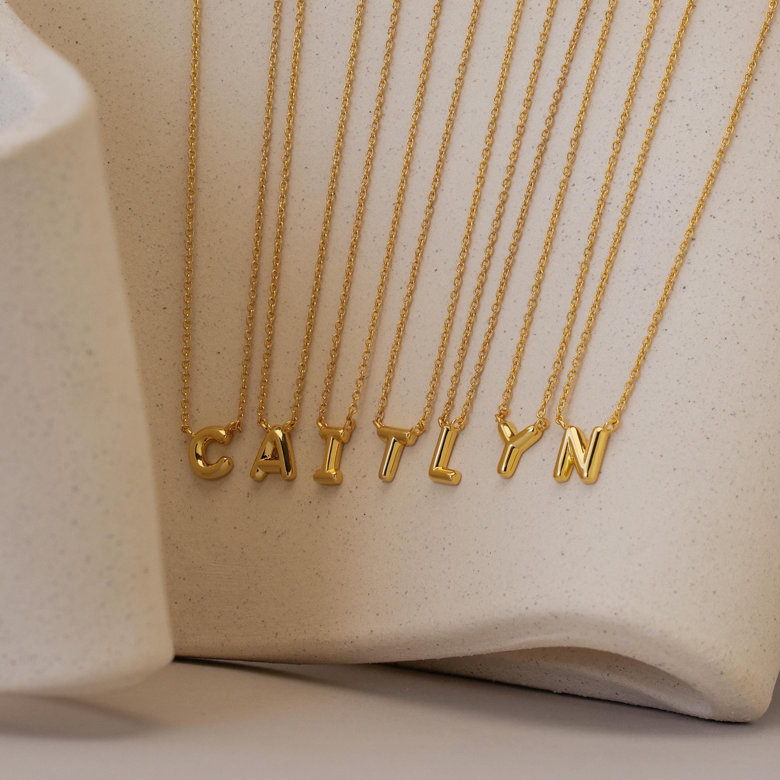 The Dainty Bubble Letter Necklace, featuring gold individual letter pendants that spell CAITLYN, is displayed on a neutral textured background for a chic and personalized accessory touch.