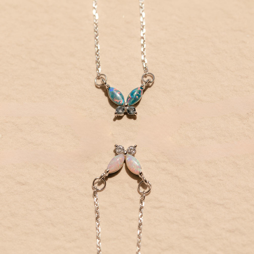 Two Opal Butterfly Necklace Sets with colored gemstone wings are displayed on a beige textured background.