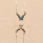 Two Opal Butterfly Necklace Sets with colored gemstone wings are displayed on a beige textured background.