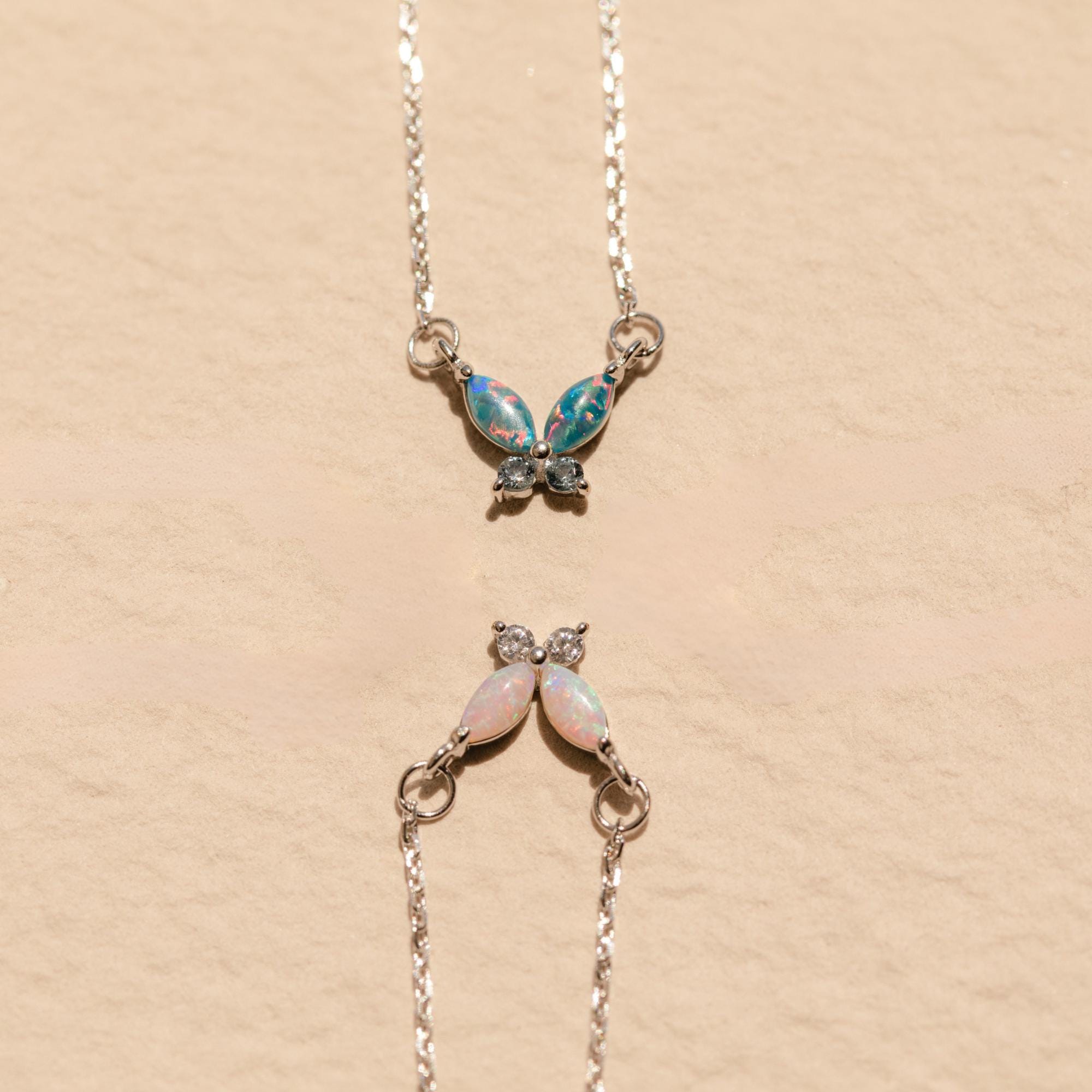 Two Opal Butterfly Necklace Sets with colored gemstone wings are displayed on a beige textured background.