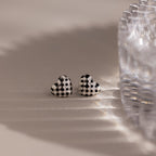 The Checkered Heart Studs are two retro-inspired earrings with a black and white checkered pattern, shown next to a glass object on a light surface.
