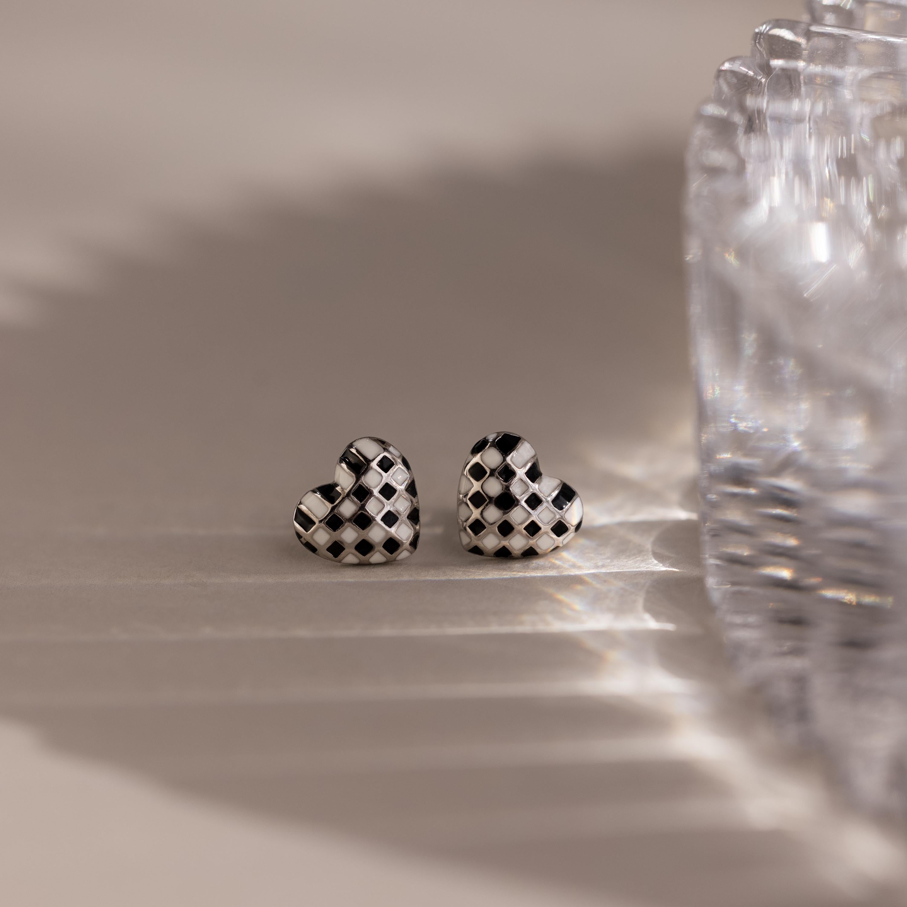 The Checkered Heart Studs are two retro-inspired earrings with a black and white checkered pattern, shown next to a glass object on a light surface.