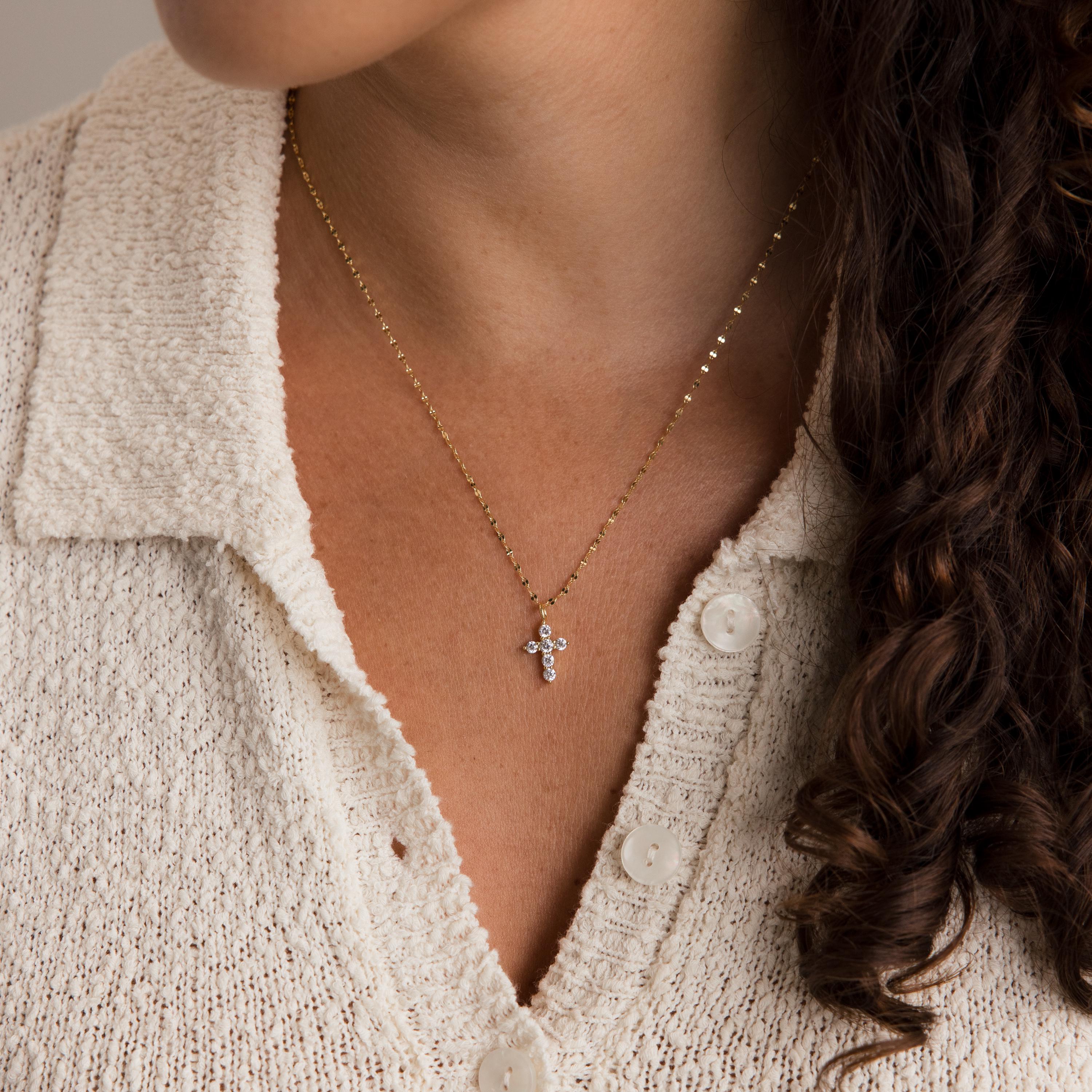 A woman wears the Diamond Cross Necklace—a gold faith necklace with pavé CZ diamonds and a small cross pendant—paired with a cream knit top; focus is on her neck and collar.
