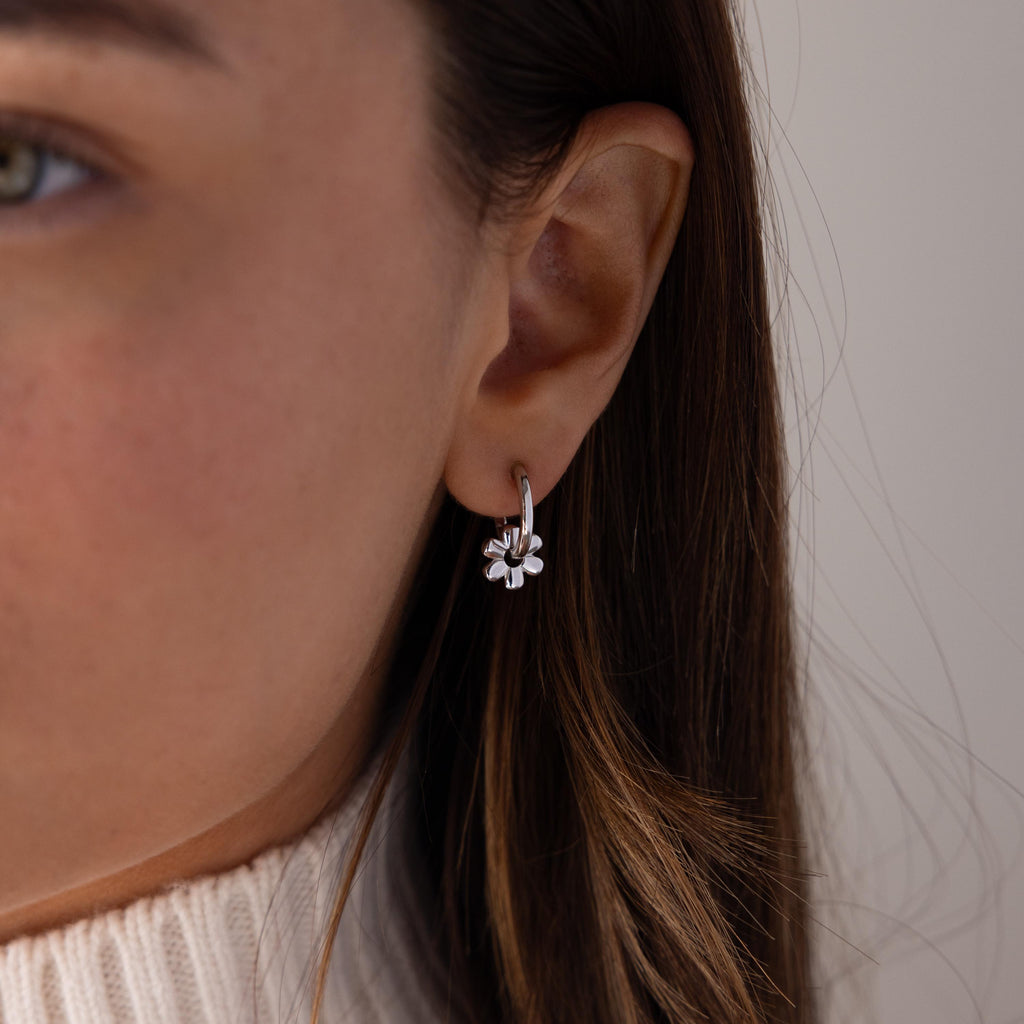 Close-up of a woman wearing Dangling Blossom Huggies, styled as silver flower charm earrings, paired with a cream turtleneck sweater.