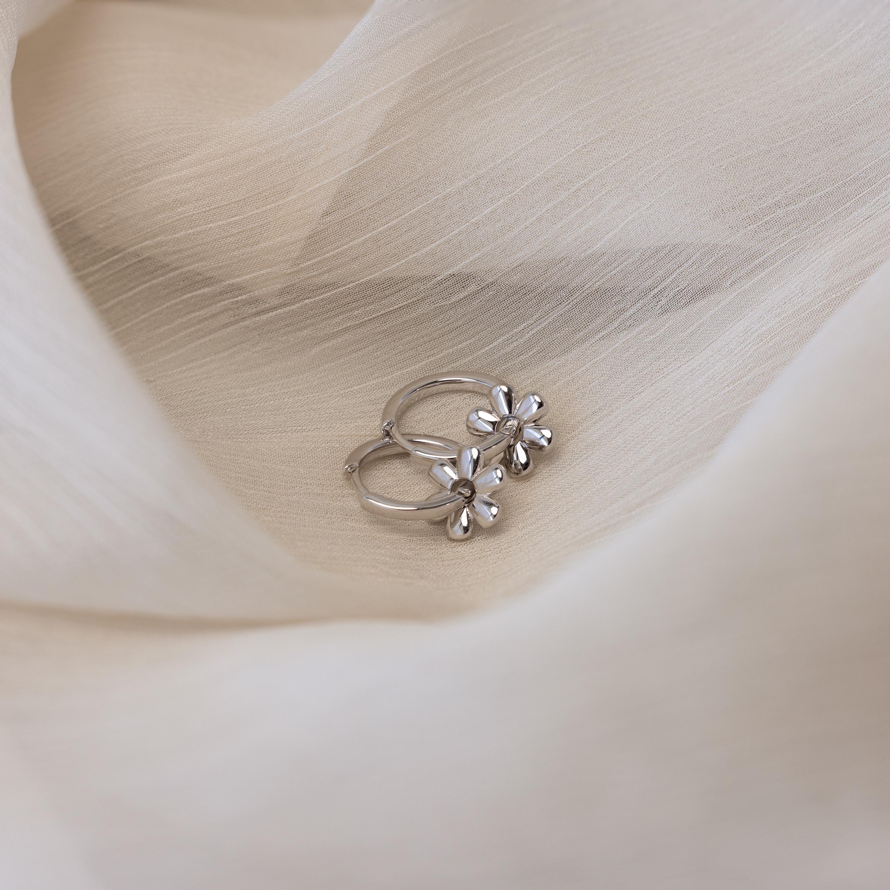 Dangling Blossom Huggies feature delicate flower charms and rest on soft beige fabric, adding elegant style to any outfit.