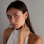 Woman with long brown hair wears Dangling Blossom Huggies and a white sleeveless knit top, gazing at the camera.