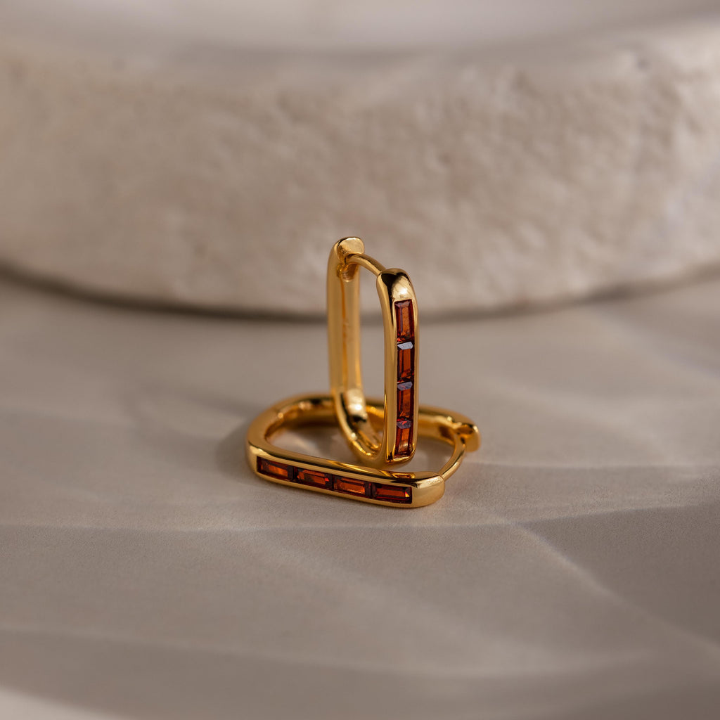 Baguette Birthstone Link Earrings: gold geometric hoops with rectangular red stones, displayed on a smooth beige surface beside a textured white stone.