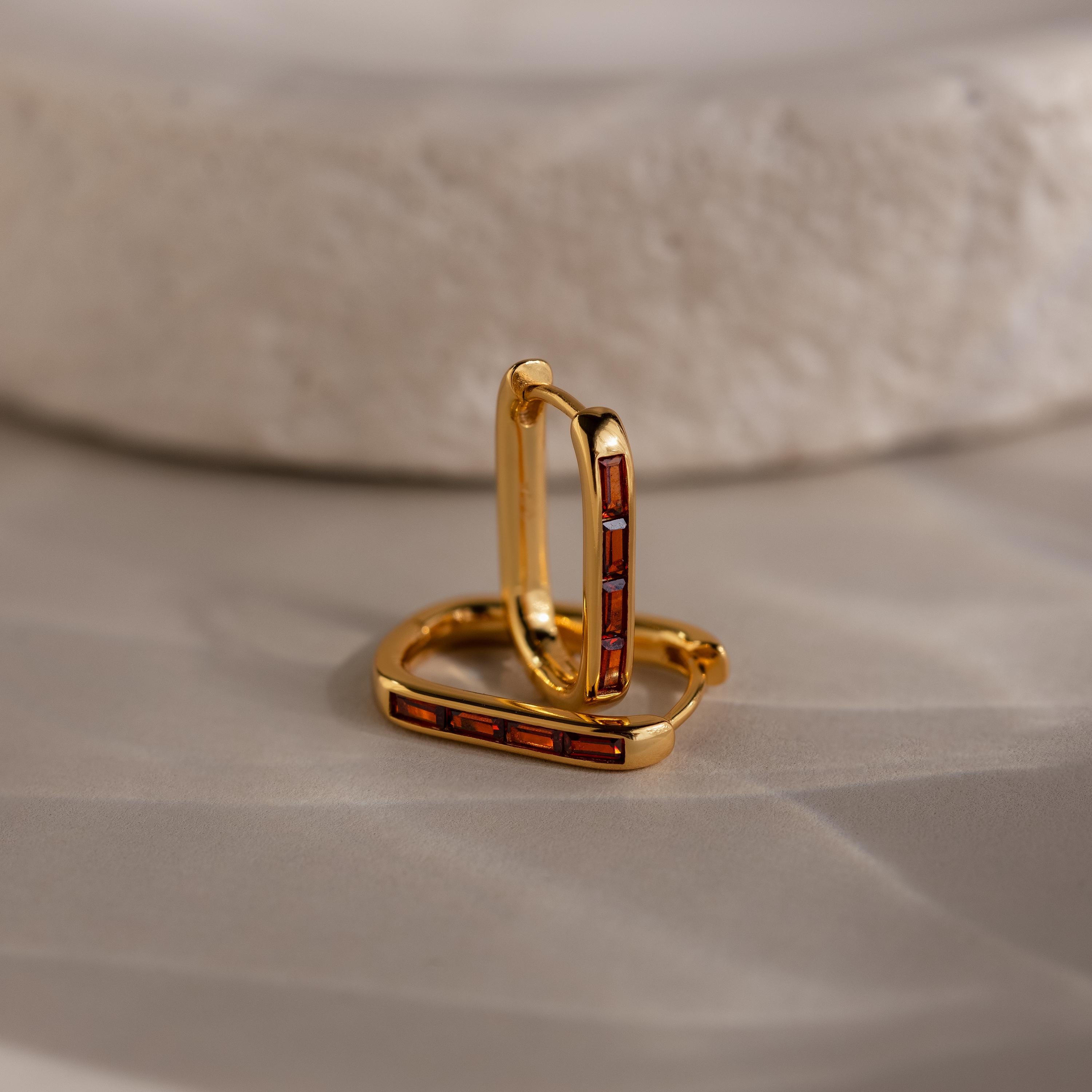 Baguette Birthstone Link Earrings: gold geometric hoops with rectangular red stones, displayed on a smooth beige surface beside a textured white stone.