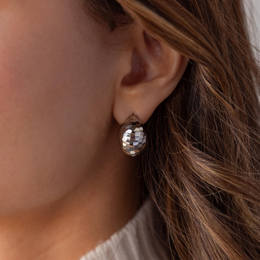 Close-up of a woman's ear wearing Bold Disco Earrings—large, faceted silver studs inspired by retro glam—with brown wavy hair visible.