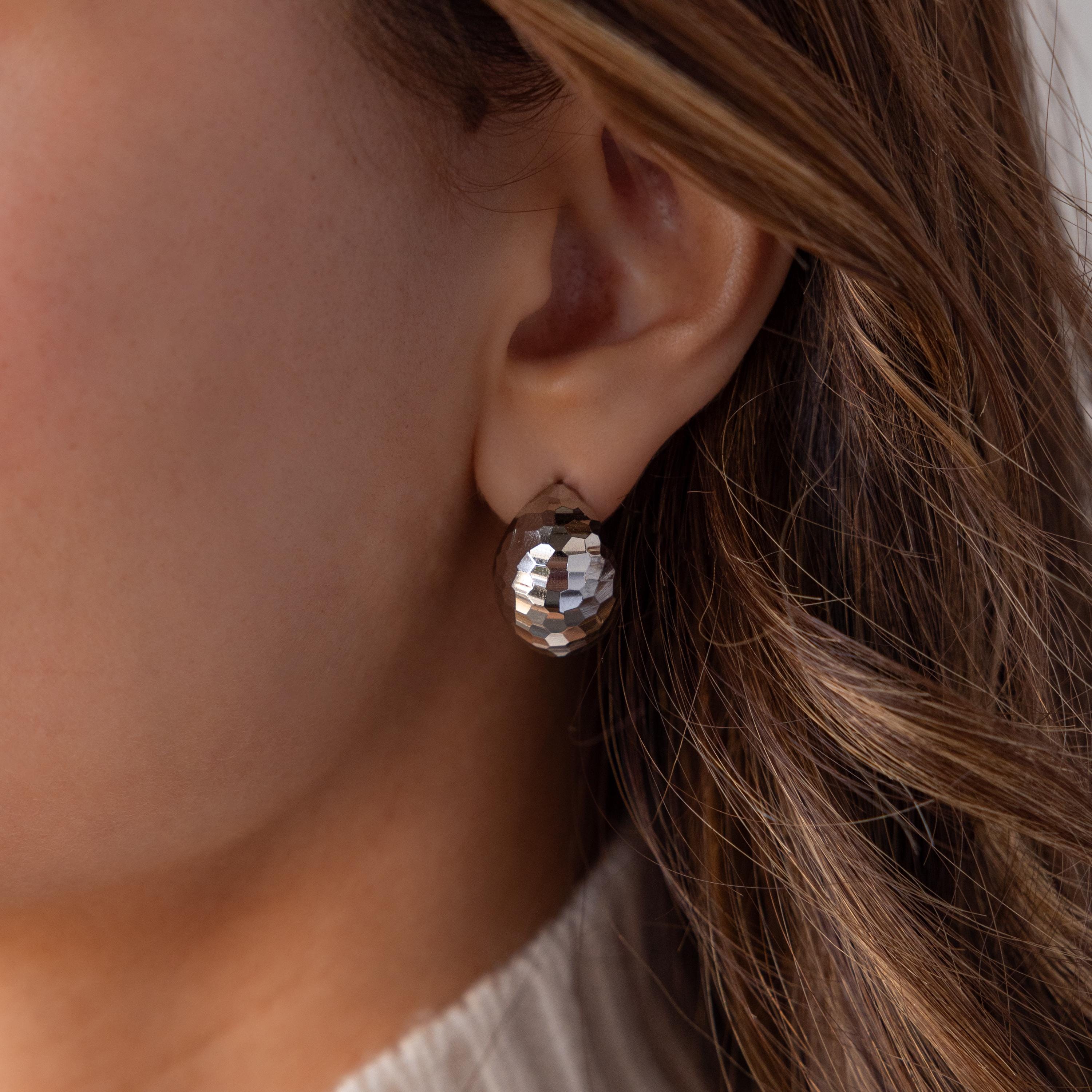 Close-up of a woman's ear wearing Bold Disco Earrings—large, faceted silver studs inspired by retro glam—with brown wavy hair visible.